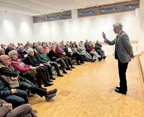 Ein Raum mit im halbkreis aufgestellten Stühlen für die Zuhörenden. Der Redner steht auf der rechten Seite und gestikuliert mit der Hand. 