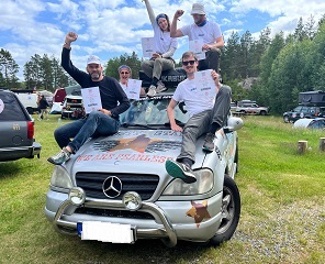 Fünf Menschen sitzen auf einem silbernen Mercedes und halten jeder Stolz eine Urkunde in die Höhe. Manche recken noch eine gestreckte Faust in den Himmel.