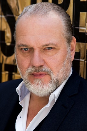 Jan-Gregor_Kremp_Schirmherr_von_PalliLev Der Schauspieler Jan-Gregor Kremp steht vor einem goldenen Hintergrund mit scharzer Schrift und guckt ernst in die Kamera.